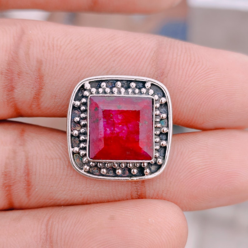 May include: A square-cut vibrant red gemstone ring set in a silver frame. The metal setting features a dark, oxidised finish with a decorative border of small, raised silver beads surrounding the central stone in a square, rounded-corner shape.