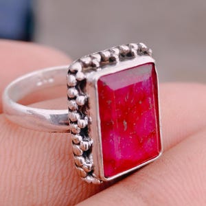 May include: A silver ring featuring a rectangular, faceted, vibrant pink gemstone set in a bezel with a decorative, beaded, or granulated metal border. The band is smooth and polished silver, showcasing a clear 925 hallmark stamp on the inner surface.
