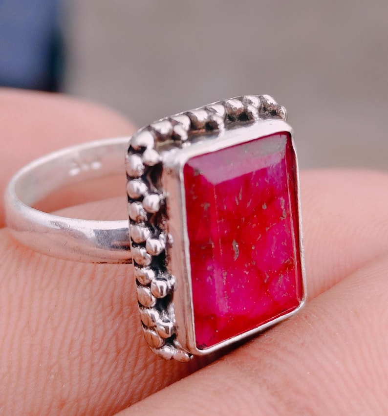 May include: A silver ring featuring a rectangular, faceted, vibrant pink gemstone set in a bezel with a decorative, beaded, or granulated metal border. The band is smooth and polished silver, showcasing a clear 925 hallmark stamp on the inner surface.