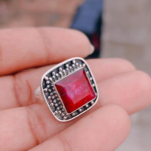 May include: A silver ring featuring a square-cut, deep red gemstone set in a frame decorated with small, raised silver beads. The metal has a darkened, oxidised finish in the recessed areas, highlighting the textured, beaded border design.