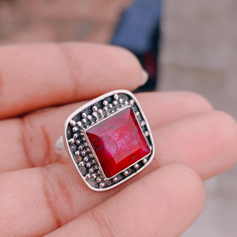 May include: A silver ring featuring a square-cut, deep red gemstone set in a frame decorated with small, raised silver beads. The metal has a darkened, oxidised finish in the recessed areas, highlighting the textured, beaded border design.