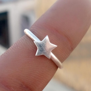 May include: A silver-toned ring featuring a small, five-pointed star charm centred on a thin, smooth band. The metal has a matte, slightly textured finish, and the star is positioned flat against the band, creating a simple, uniform appearance.