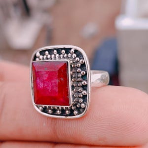 May include: A silver ring featuring a square-cut, deep red gemstone set in a frame decorated with small metal beads. The setting has an oxidised finish, and the band is smooth and polished, showcasing a clear contrast between the red stone and metal.