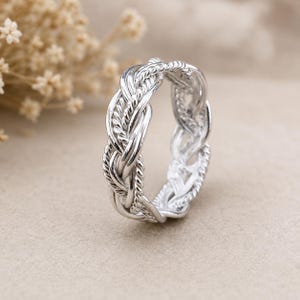 May include: A silver ring featuring a complex braided design composed of multiple twisted metal strands. The band has a polished, reflective finish and an intricate, woven pattern that wraps continuously around the entire circumference of the ring.