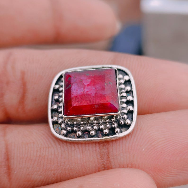 May include: A square-cut, deep red gemstone set in a silver ring. The setting features a raised, textured border with small, rounded silver beads surrounding the central stone. The metal has a darkened, oxidised finish in the recessed areas.
