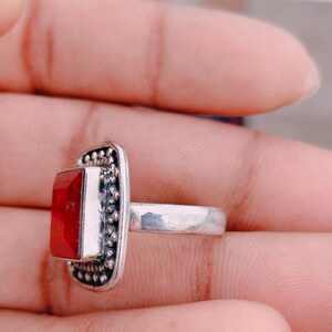 May include: A silver ring featuring a rectangular, faceted red stone set in a frame decorated with small metal beads. The band is smooth and metallic, and the setting has a textured, beaded border surrounding the central red gemstone.