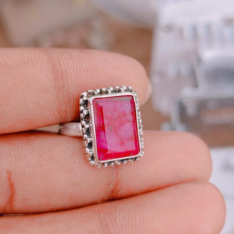 May include: A silver ring featuring a rectangular, faceted, vibrant pinkish-red gemstone set in a textured, beaded metal frame. The band is smooth and metallic, showcasing a clear contrast between the polished silver setting and the deep red stone.