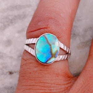 Puede incluir: Un anillo con una incrustación ovalada de concha de abulón iridiscente en tonos azules y verdes vibrantes. La banda está hecha de un material metálico con una superficie texturizada y muescas que se divide en dos segmentos cerca de la piedra.