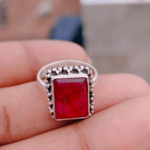 May include: A silver ring featuring a rectangular, faceted deep red gemstone set in a frame with a beaded, textured border. The metal band is smooth and polished, supporting the prominent, vertically oriented stone setting.