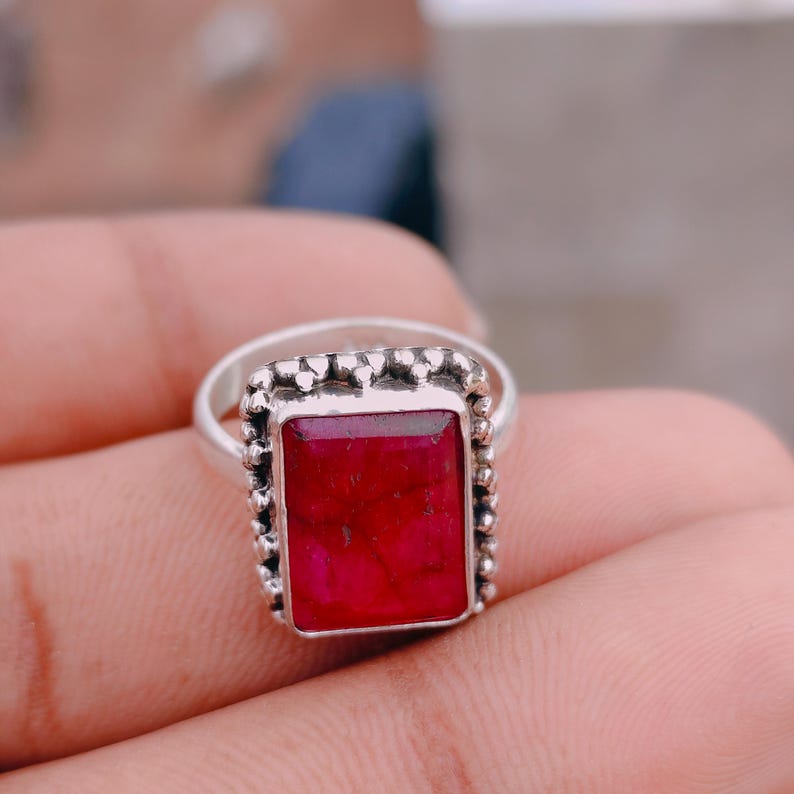 May include: A silver ring featuring a rectangular, faceted deep red gemstone set in a frame with a beaded, textured border. The metal band is smooth and polished, supporting the prominent, vertically oriented stone setting.