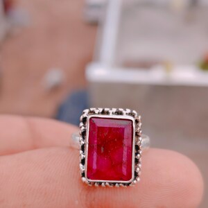 May include: A silver ring featuring a rectangular, deep red gemstone with visible internal fractures. The stone is set in a decorative, beaded silver bezel frame that surrounds the entire perimeter of the rectangular setting, mounted on a plain band.