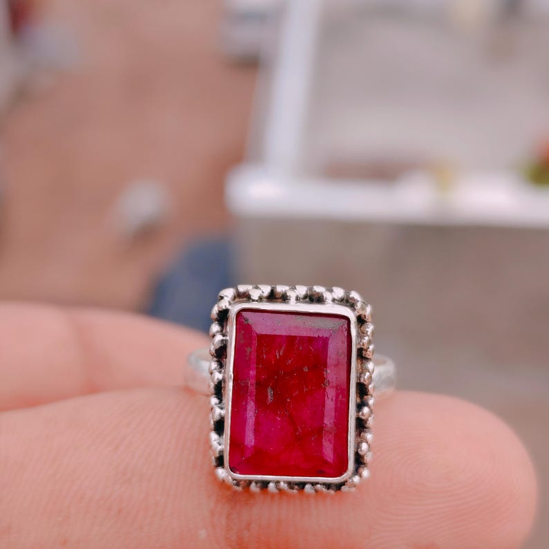 May include: A silver ring featuring a rectangular, deep red gemstone with visible internal fractures. The stone is set in a decorative, beaded silver bezel frame that surrounds the entire perimeter of the rectangular setting, mounted on a plain band.