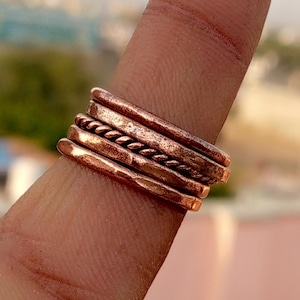 May include: A stack of copper-colored rings, each with a unique texture, including a twisted rope design. The rings are worn on a finger, showcasing their varying widths and finishes. The overall aesthetic is rustic and handcrafted.