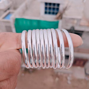 Può includere: Un set di nove braccialetti rigidi color argento lucido con una superficie liscia e arrotondata. Ogni braccialetto presenta un design circolare semplice e continuo con una finitura metallica riflettente, disposti fianco a fianco.
