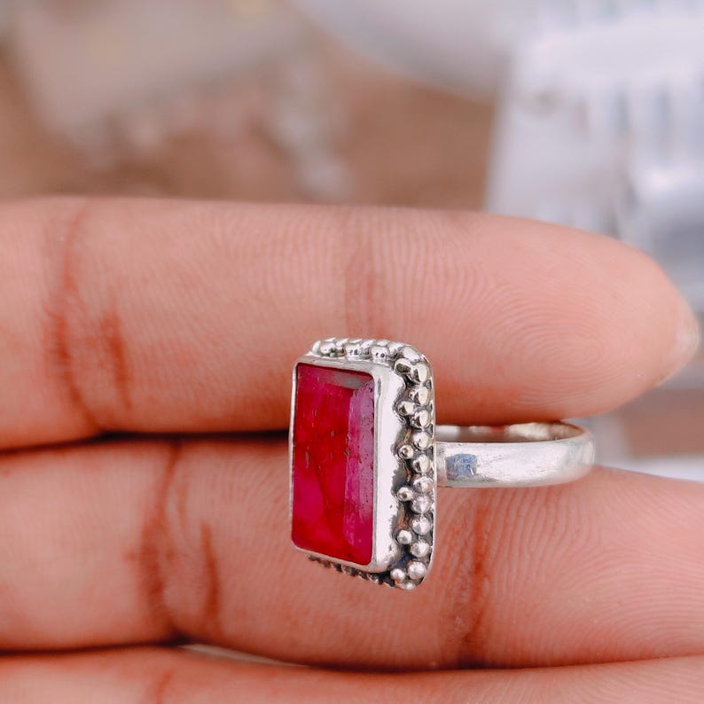 May include: A silver ring featuring a rectangular, deep red gemstone set in a bezel. The metal surrounding the stone is decorated with a series of small, raised silver spheres, and the band is a smooth, polished silver metal finish.