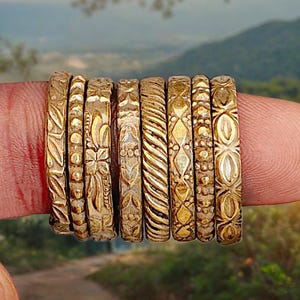 May include: A collection of gold-colored rings with intricate embossed designs, including floral, geometric, and textured patterns. The rings are displayed on a finger, showcasing their varied details. The background features a blurred landscape.