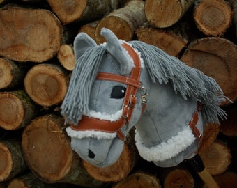 May include: A gray and white stuffed horse head with a brown leather bridle. The horse is posed against a background of stacked wooden logs.