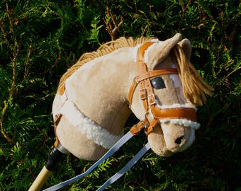 May include: A tan hobby horse with a light brown mane and a brown leather bridle. The toy horse has a white fluffy trim and blue reins. The background is a green bush.