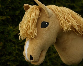 May include: A plush toy horse head in shades of tan and cream, with a flowing mane and a white stripe on its face. The horse has a black eye and a black nostril, set against a blurred green background.