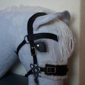May include: A soft white horse head with a black leather bridle. The bridle has silver-coloured buckles and decorative details. The horse has a white mane and a grey-tipped ear.