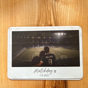 Könnte beinhalten: Ein rechteckiger Fotodruck mit einer Stadionansicht und zwei Personen auf der Tribüne. Eine Person trägt ein dunkles Trikot mit dem Namen MAJEWSKI und der Nummer 8. Unter dem Bild steht der Text Matchday mit einem Herz und 1.9.2026.