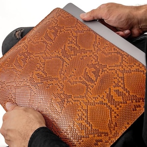May include: A brown leather laptop sleeve with a snakeskin pattern. The sleeve is being held by a person's hands.