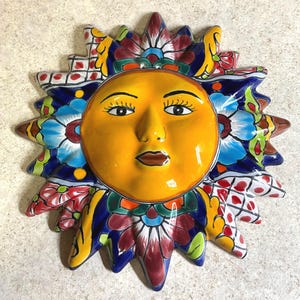 May include: A ceramic sun wall plaque featuring a central yellow face with dark eyes and lips. The surrounding rays are decorated with intricate, colorful floral patterns in shades of blue, red, green, and yellow, creating a vibrant, multi-colored design.