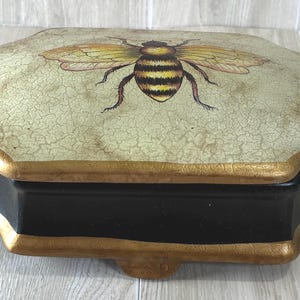 May include: A decorative box with a black base and gold-painted edges. The lid features a cream-colored surface with a crackled texture and a central illustration of a yellow and black bee with translucent wings, showcasing a detailed insect design.