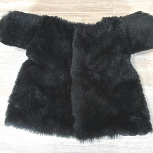 May include: A black faux fur jacket with short sleeves. The jacket is open in the front and has a soft, fluffy texture. The garment is laid flat on a light-colored wooden surface.