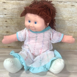 May include: A vintage doll with a soft, plush body. The doll has brown yarn hair, blue eyes, and freckles. It wears a light pink and white star-patterned dress with blue trim and white shoes. The doll's arms are outstretched.