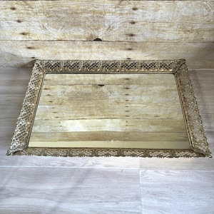 May include: A rectangular, antique-style mirror with an ornate, gold-colored filigree frame. The mirror's surface reflects the surrounding environment. The frame features a detailed, openwork design, adding a vintage aesthetic.
