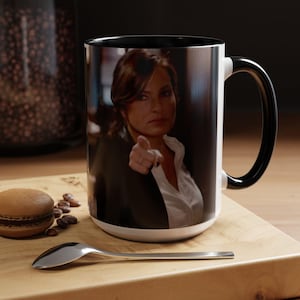 Puede incluir: Taza de cerámica blanca con interior y asa negros. La taza presenta una imagen a color de una persona señalando. La taza está sobre una superficie de madera con una cuchara, granos de café y un macaron.