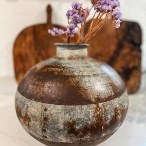 Könnte beinhalten: Eine runde, rostige Metallvase mit einem braunen Rostband. Die Vase enthält getrocknete, lila Blüten. Die Vase hat einen Vintage-Look, geeignet für die Inneneinrichtung. Der Vasendurchmesser beträgt ca. 20 cm.