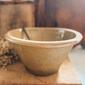 May include: A tan-colored ceramic mixing bowl with a wide, rounded rim and a smooth, glazed interior surface. The bowl features a tapered, conical shape that narrows toward the base, showcasing a consistent earthy tone throughout the exterior.