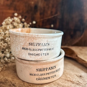 May include: Two antique, off-white ceramic pots stacked on top of each other. The pots have a cracked, aged appearance and feature the text "SHIPPAM'S HIGH CLASS POTTED MEAT CHICHESTER" in black lettering. The background is a rustic wooden surface.