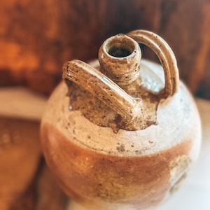 Op de afbeelding: Een rustieke, ronde keramische kruik met een handvat en een kleine opening. De kruik heeft een crèmekleurige basis met een roodbruine band in het midden. Het handvat en de opening zijn donkerder bruin, wat het een vintage uitstraling geeft.