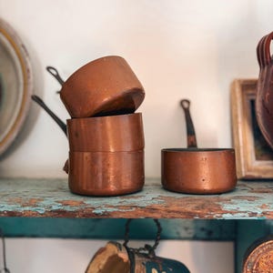 Op de afbeelding: Een verzameling antieke koperen steelpannen met zwarte handvatten, gestapeld op een verweerde turquoise plank. Het verouderde metalen kookgerei heeft een warme, roodbruine tint, wat wijst op vintage keukengerei.