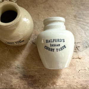 May include: Two antique ceramic jars with a cream-colored glaze. One jar has the text "Green label" and the other reads "HALFORD'S INDIAN CURRY PASTE" in dark blue lettering. The jars sit on a wooden surface.