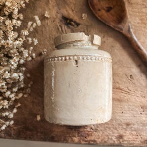 Puede incluir: Un pequeño tarro de cerámica beige antiguo con cuerpo cilíndrico y una banda decorativa de cuentas alrededor de la parte superior. El tarro tiene una tapa con una pequeña muesca. Una cuchara de madera y flores secas están en el fondo.