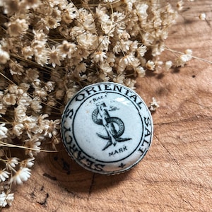 May include: A circular, white ceramic lid featuring black text and a mermaid design. The text reads "ORIENTAL SALT" and "SEARCH'S TRACE MARK". The lid is placed on a wooden surface, with dried flowers in the background.