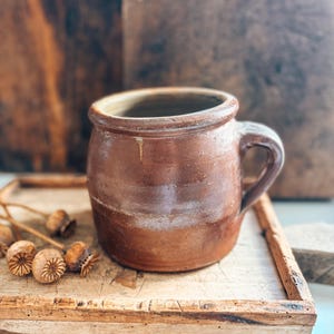 May include: A rustic, brown ceramic mug with a handle. The mug has a slightly uneven surface and a dark brown base. It sits on a wooden tray with dried seed pods.