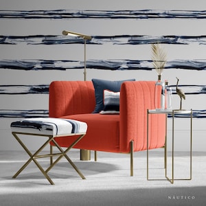 May include: A coral-colored armchair with gold legs and a matching ottoman. The armchair has a tufted back and a white and blue patterned cushion. The ottoman has a white and blue patterned top. The armchair and ottoman are in front of a white wall with blue and gray horizontal stripes.