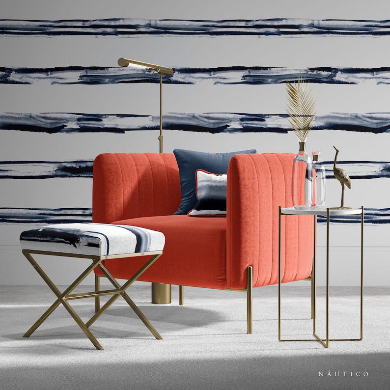 May include: A coral-colored armchair with gold legs and a matching ottoman. The armchair has a tufted back and a white and blue patterned cushion. The ottoman has a white and blue patterned top. The armchair and ottoman are in front of a white wall with blue and gray horizontal stripes.