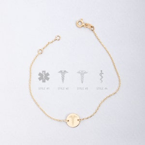 Op de afbeelding: Een gouden kettingarmband met een ronde bedel met een medisch symbool. De bedel heeft een caduceus-ontwerp met een slang die om een staf met vleugels is gewikkeld. De armband wordt getoond tegen een witte achtergrond.