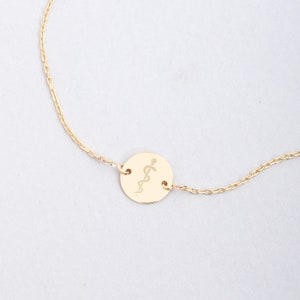 Op de afbeelding: Een gouden ketting met een ronde hanger met een gegraveerd medisch symbool, de staf van Asklepios.