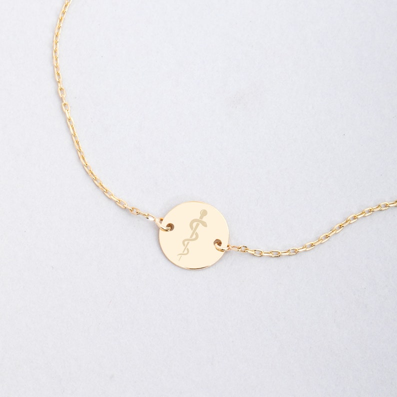 Op de afbeelding: Een gouden ketting met een ronde hanger met een gegraveerd medisch symbool, de staf van Asklepios.