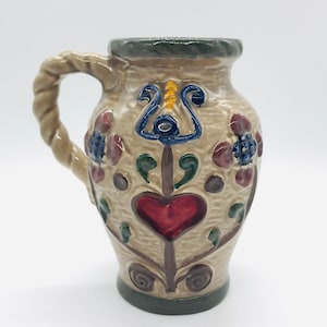 May include: A small, beige ceramic pitcher with a twisted handle. The pitcher is decorated with colorful floral designs, including red, blue, green, and yellow. The pitcher has a heart-shaped design in the center.