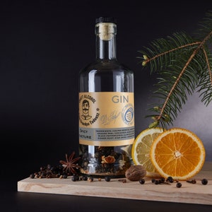 Puede incluir: Una botella de cristal transparente de ginebra con tapón de corcho, etiquetada como "Grandpa Tadeusz Spicy Tincture". La botella está rodeada de bayas de enebro, anís estrellado y rodajas de cítricos sobre una tabla de madera. Una ramita de pino está en el fondo.
