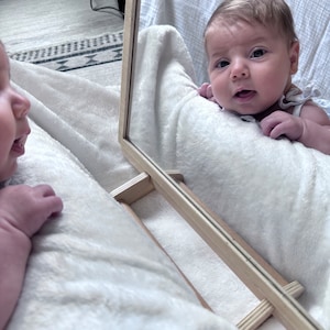 May include: A baby looking into a mirror, smiling and reaching out with their hand. The baby is lying on a white blanket.