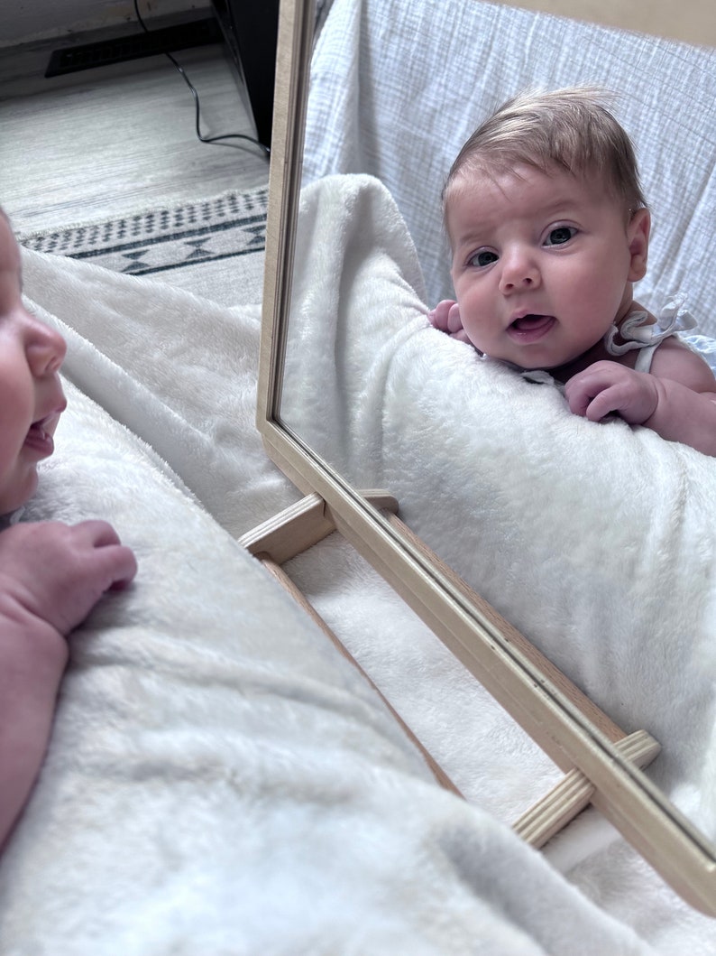 May include: A baby looking into a mirror, smiling and reaching out with their hand. The baby is lying on a white blanket.